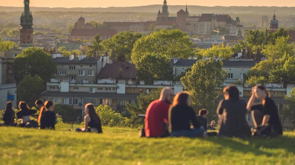 People sitting on a grassy hill overlooking Krakow's rooftops and Wawel Castle at sunset